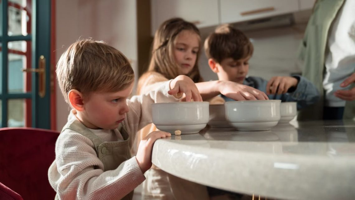 Three children gather around a table enjoying breakfast indoors on a sunny morning.