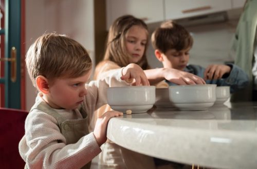 Three children gather around a table enjoying breakfast indoors on a sunny morning.