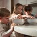 Three children gather around a table enjoying breakfast indoors on a sunny morning.